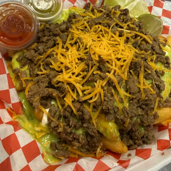 Loaded fries with ground beef, melted cheese, green sauce, and two lime wedges, served in a red and white checkered paper.