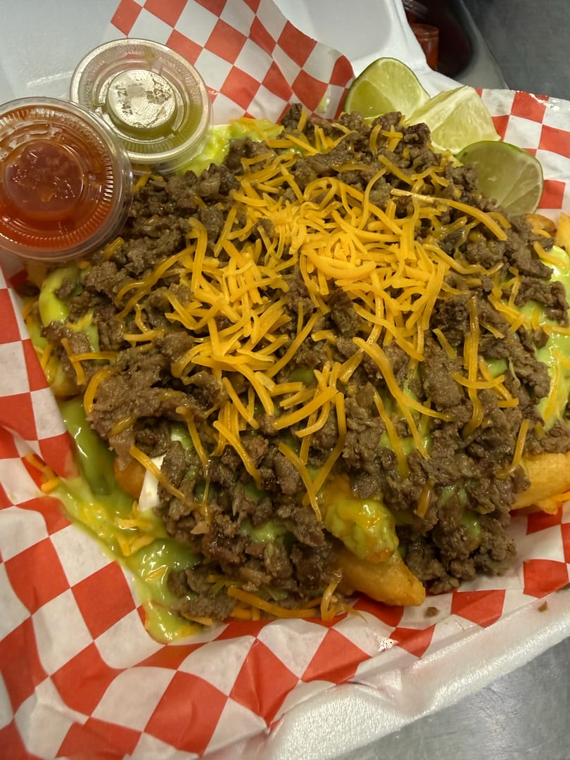 Loaded fries with ground beef, melted cheese, green sauce, and two lime wedges, served in a red and white checkered paper.