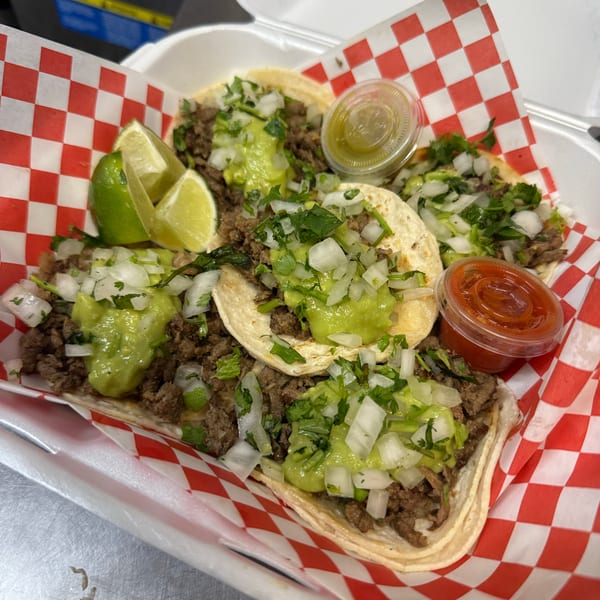Four street tacos with meat, guacamole, onions, cilantro, lime wedges, and two sauces in a takeout container.