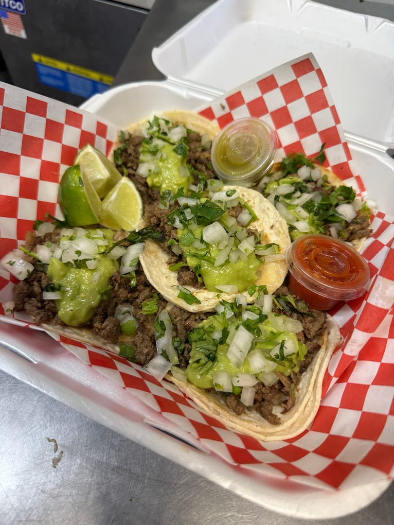 Four street tacos with meat, guacamole, onions, cilantro, lime wedges, and two sauces in a takeout container.
